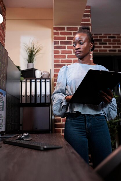 Investment company african american professional financial analyst with clipboard analyzing real time market data. Brokerage agency agent standing in front of multi monitor workstation with clipboard.