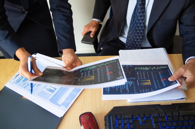 Инвестор и трейдер обсуждают статистические данные, держат в руках бумаги с финансовыми диаграммами и ручкой. Обрезанный снимок. Работа брокера или торговая концепция