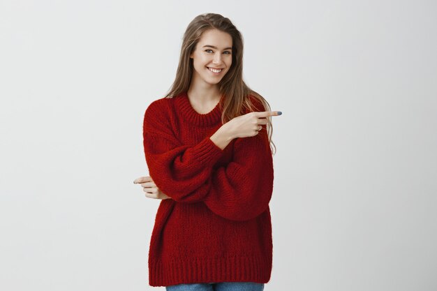 Inviting friends for cup of tea. Portrait of confident good-looking caucasian female freelancer in loose sweater, pointing right with index finger and smiling with approval, liking idea over gray wall