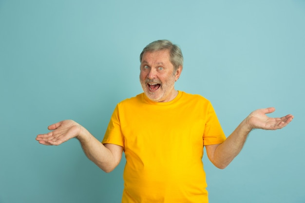 Inviting, uncertain gesture. Caucasian man portrait isolated on blue studio background. Beautiful male model in yellow shirt posing. Concept of human emotions, facial expression, sales, ad. Copyspace.