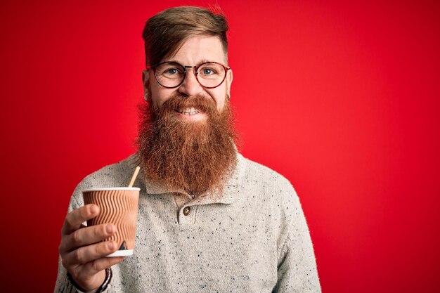 Ирландский рыжий мужчина с бородой пьет кофе на вынос из бумажного стаканчика на красном фоне со счастливым лицом, стоящим и улыбающимся с уверенной улыбкой, показывающей зубы