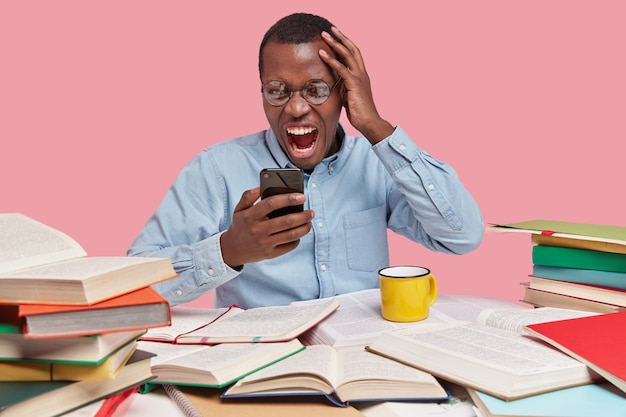 Free photo irked angered dark skinned young man looks with discontent peevish facial expression at cell phone, dressed in formal shirt, keeps hand on head