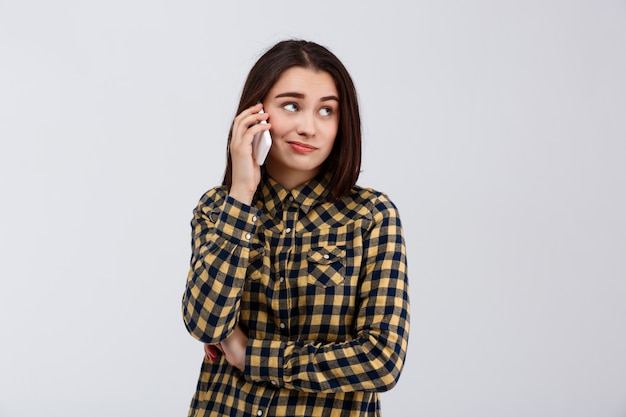 Ироничная молодая красивая девушка одета в клетчатой рубашке, выступая на телефоне, глядя на стороне над белой стеной.