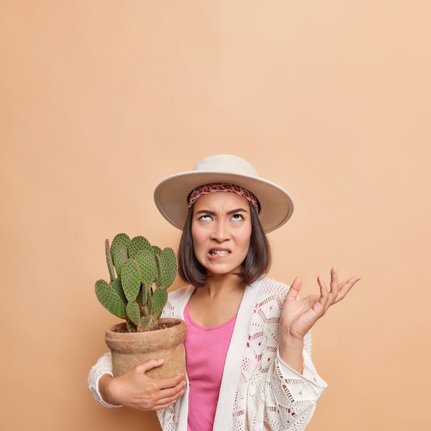 Irritated Asian lady bites lips raises palm looks angrily above holds potted cactus wears fedora white knitted coat poses against brown wall copy space for your promotion