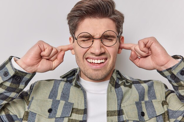 Irritated brunet adult man plugs ears with index fingers grimaces from loud noise disturbed by noisy neighbours wears round spectacles casual shirt isolated over white background. Turn sound off