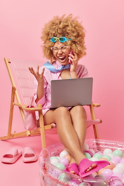 Irritated curly haired woman freelancer screams angrily makes phone call works on laptop computer dressed in summer clothes poses on deck chair angry to have spoiled holidays because of work