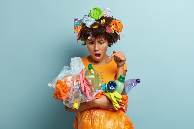 Irritated emotional dark skinned female volunteer collects plastic bottles and polythene objects, fights against nature disaster or pollution problem, gathers trash, isolated against blue wall