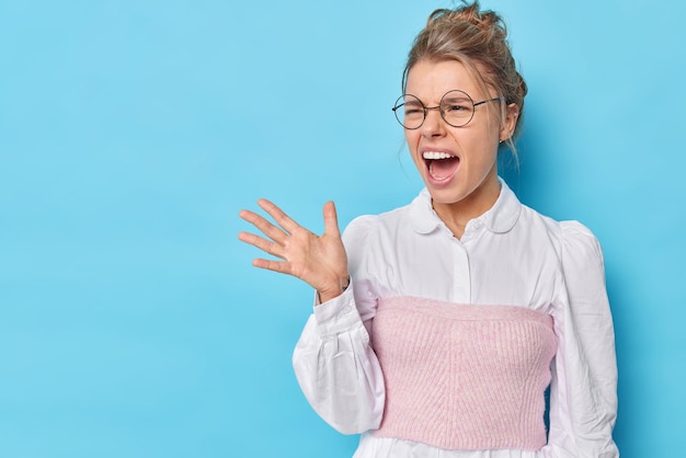 Foto gratuita insegnante donna irritata urla con rabbia ai gesti degli alunni e tiene la bocca aperta indossa occhiali rotondi e camicia bianca ordinata isolata sul blu con spazio vuoto per la copia. emozioni negative