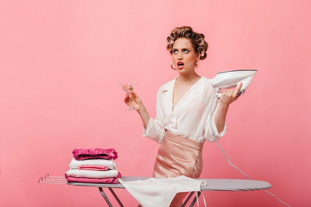 Irritated housewife in beautiful outfit holding martini glass and ironing clothes on pink wall