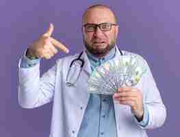 Free photo irritated middle-aged male doctor wearing medical robe and stethoscope with glasses holding and pointing at money  isolated on purple wall