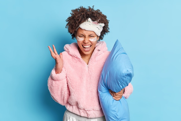 Irritated millennial girl raises hand and exclaims angrily being annoyed with noisy neighbours who interrupt to sleep poses in pajama holds soft pillow isolated on blue wall