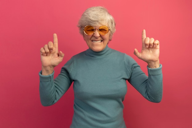 Irritated old woman wearing blue turtleneck sweater and sunglasses biting lip pointing up with closed eyes isolated on pink wall