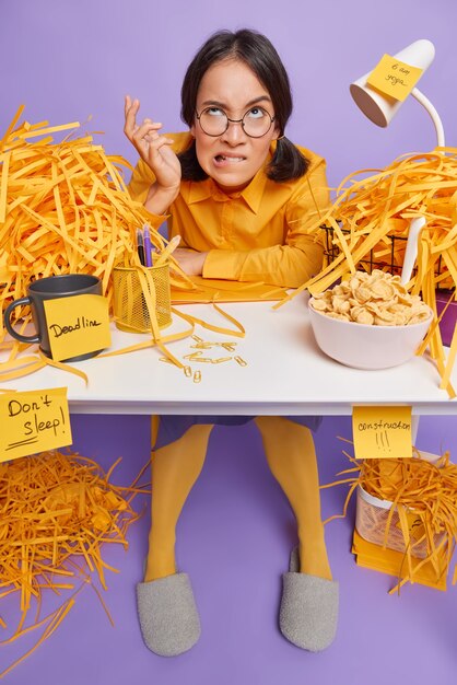 Free photo irritated puzzled schoolgirl bites lips has much work to do sits alone at desktop has deadline to prepare for test has cereals for breakfast surrounded by cut papers isolated over purple wall