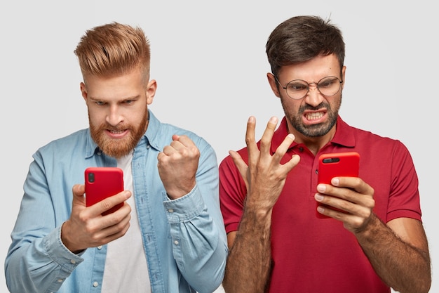 Free photo irritated two guys look angrily at screen of smart phones, watch football match online, being annoyed as favourite team lost game, focused at something, dressed in fashionable clothes, pose indoor