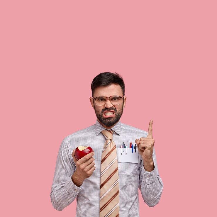 मुफ़्त फोटो irritated unshaven entrepreneur points at blank space above, frowns face in discontent, wears big spectacles, eats red apple