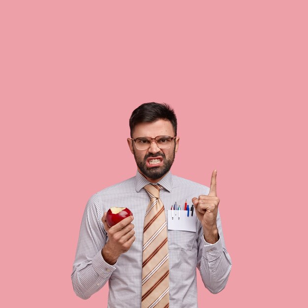 Irritated unshaven entrepreneur points at blank space above, frowns face in discontent, wears big spectacles, eats red apple