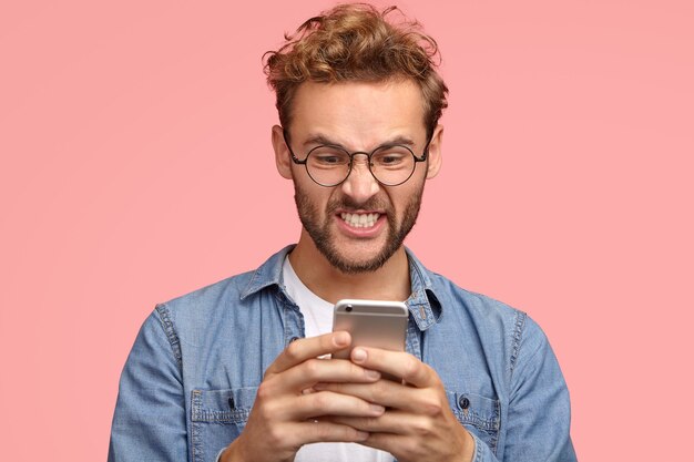 Irritated unshaven man has peevish facial expression, holds modern cell phone in hands