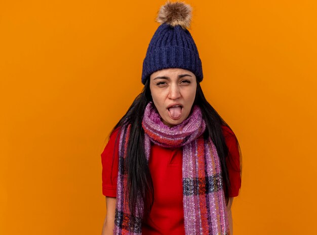 복사 공간 오렌지 벽에 고립 된 혀를 보여주는 겨울 모자와 스카프를 착용 초조하고 약한 젊은 백인 아픈 소녀