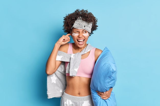 Irritated young Afro American woman plugs ears cannot sleep in such noisy place looks angrily aside exclaims and asks not to bother her holds pillow wears pajama poses against blue wall