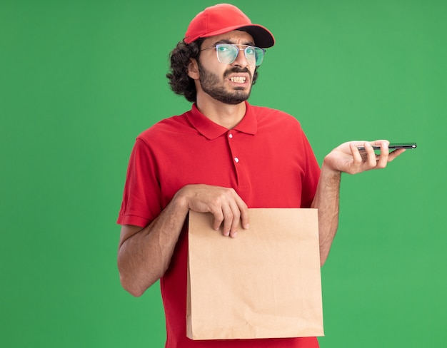 Irritato giovane fattorino caucasico in uniforme rossa e berretto con gli occhiali che tengono in mano un pacchetto di carta e un telefono cellulare