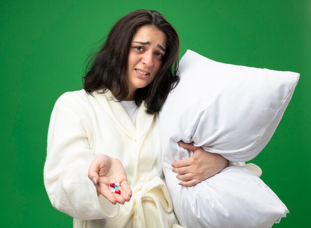 Раздраженная молодая кавказская больная девушка в халате держит подушку, протягивая медицинские капсулы к камере, глядя в камеру, изолированную на зеленом фоне