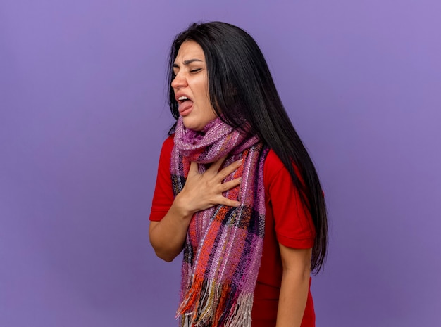Irritated young caucasian ill girl wearing scarf standing in profile view putting hand on chest showing tongue with closed eyes isolated on purple wall with copy space