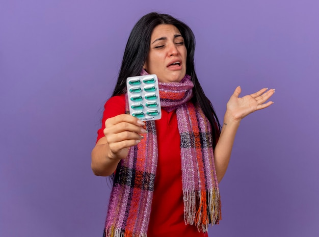 Irritated young caucasian ill girl wearing scarf stretching out pack of capsules  showing empty hand with closed eyes isolated on purple wall with copy space