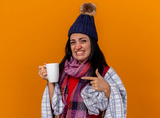 Irritated young caucasian ill girl wearing winter hat and scarf wrapped in plaid holding and pointing at cup of tea  isolated on orange wall with copy space