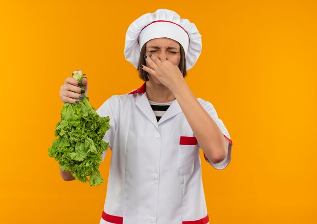 Irritated young female cook in chef uniform holding lettuce and her nose with closed eyes isolated on orange