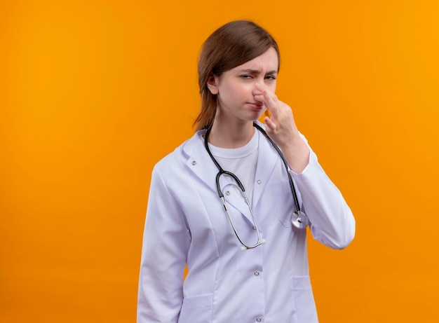 Раздраженная молодая женщина-врач в медицинском халате и стетоскопе держит нос на изолированном оранжевом пространстве с копией пространства