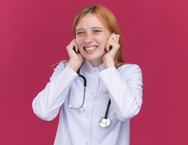 Irritated young female ginger doctor wearing medical robe and stethoscope putting fingers in ears