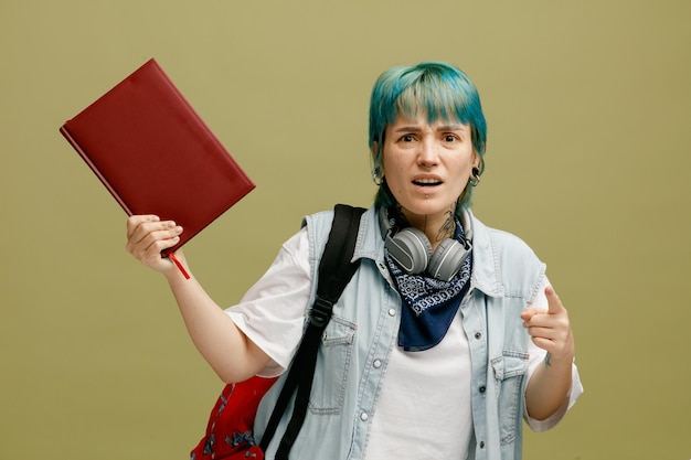 Irritated young female student wearing headphones and bandana on neck and backpack showing note book looking and pointing at camera isolated on olive green background