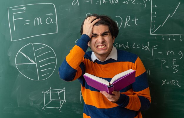 irritated young geometry teacher standing in front of chalkboard in classroom holding book looking at front keeping hand on head