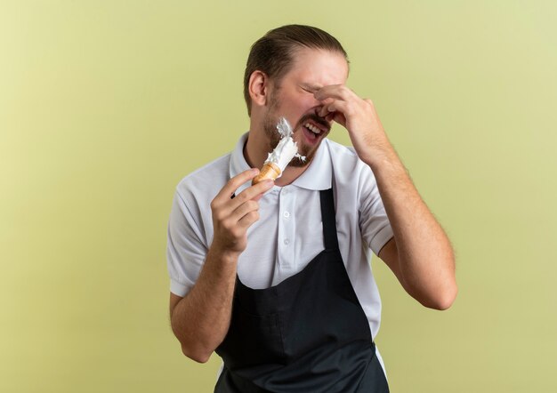 Irritated young handsome barber wearing uniform applying shaving cream on his own beard with shaving brush and holding nose with closed eyes isolated on olive green