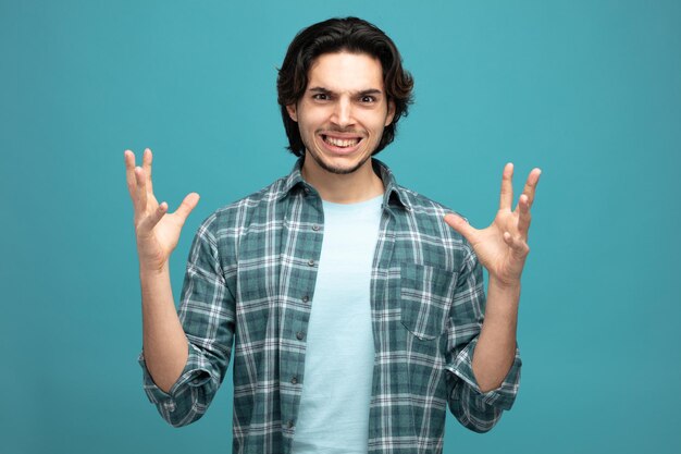 irritated young handsome man keeping hands in air looking at camera isolated on blue background