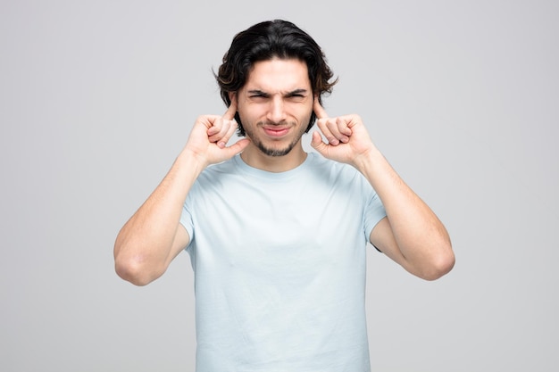 irritated young handsome man putting fingers in ears looking at camera isolated on white background