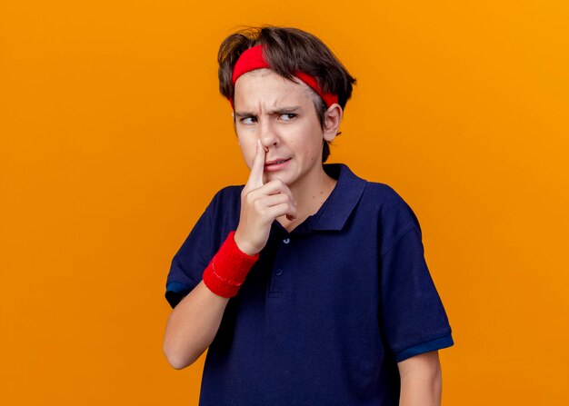 Irritated young handsome sporty boy wearing headband and wristbands with dental braces looking at side putting finger on nose isolated on orange wall with copy space