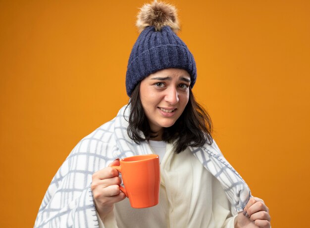 Irritated young ill woman wearing robe winter hat wrapped in plaid holding cup of tea looking at front grabbing plaid isolated on orange wall