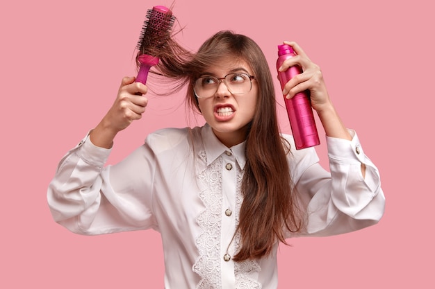 Foto gratuita la giovane donna irritata fa l'acconciatura con lacca e spazzola per capelli, ha i capelli arruffati