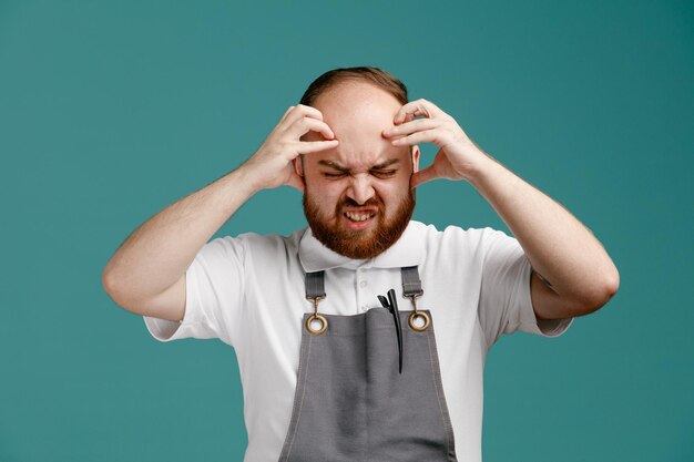 Irritated young male barber wearing white shirt and barber apron keeping hands on head with closed eyes isolated on blue background