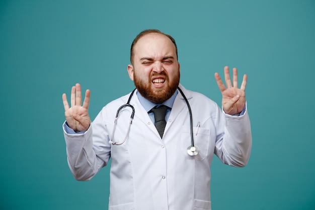 Irritated young male doctor wearing medical coat and stethoscope around his neck looking at camera showing eight with hands isolated on blue background