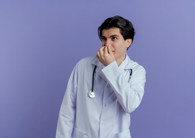 Irritated young male doctor wearing medical robe and stethoscope holding nose looking at side isolated on purple wall with copy space