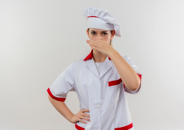 Irritated young pretty cook in chef uniform putting hand on waist and holding nose isolated on white wall