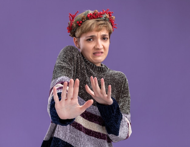 Irritated young pretty girl wearing christmas head wreath looking at camera doing refusal gesture isolated on purple background