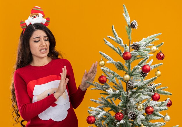 오렌지 배경에 고립 거부 제스처를하고 그것을보고 장식 된 크리스마스 트리 근처에 서있는 산타 클로스 머리띠와 스웨터를 입고 짜증이 젊은 예쁜 여자