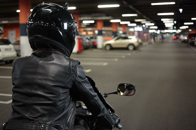 Isolated back view of female biker driving two wheeled sportbike along underground paking lot hallway, going to park her motorcycle after night ride. Motorcycling, extreme sports and lifestyle