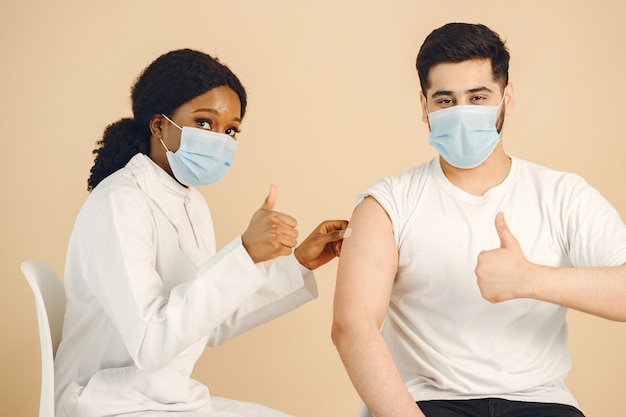 Isolated, beige background. African woman medical worker and man in masks showing thumbs up. Vaccination against covid.