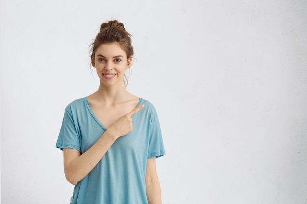 캐주얼 블루 티셔츠에 고립 된 쾌활 한 젊은 여자는 광범위하게 웃 고, 흰색 copyspace에서 그녀의 검지 손가락을 가리키는