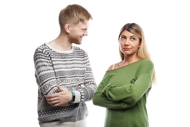 Isolated cropped shot of displeased girl in green long sleeved top keeping arms folded, feeling bored while having small talk with nerdy young bearded man with cheerful smile. People and relationships