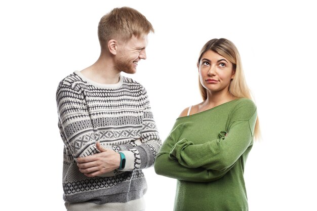 Isolated cropped shot of displeased girl in green long sleeved top keeping arms folded, feeling bored while having small talk with nerdy young bearded man with cheerful smile. People and relationships